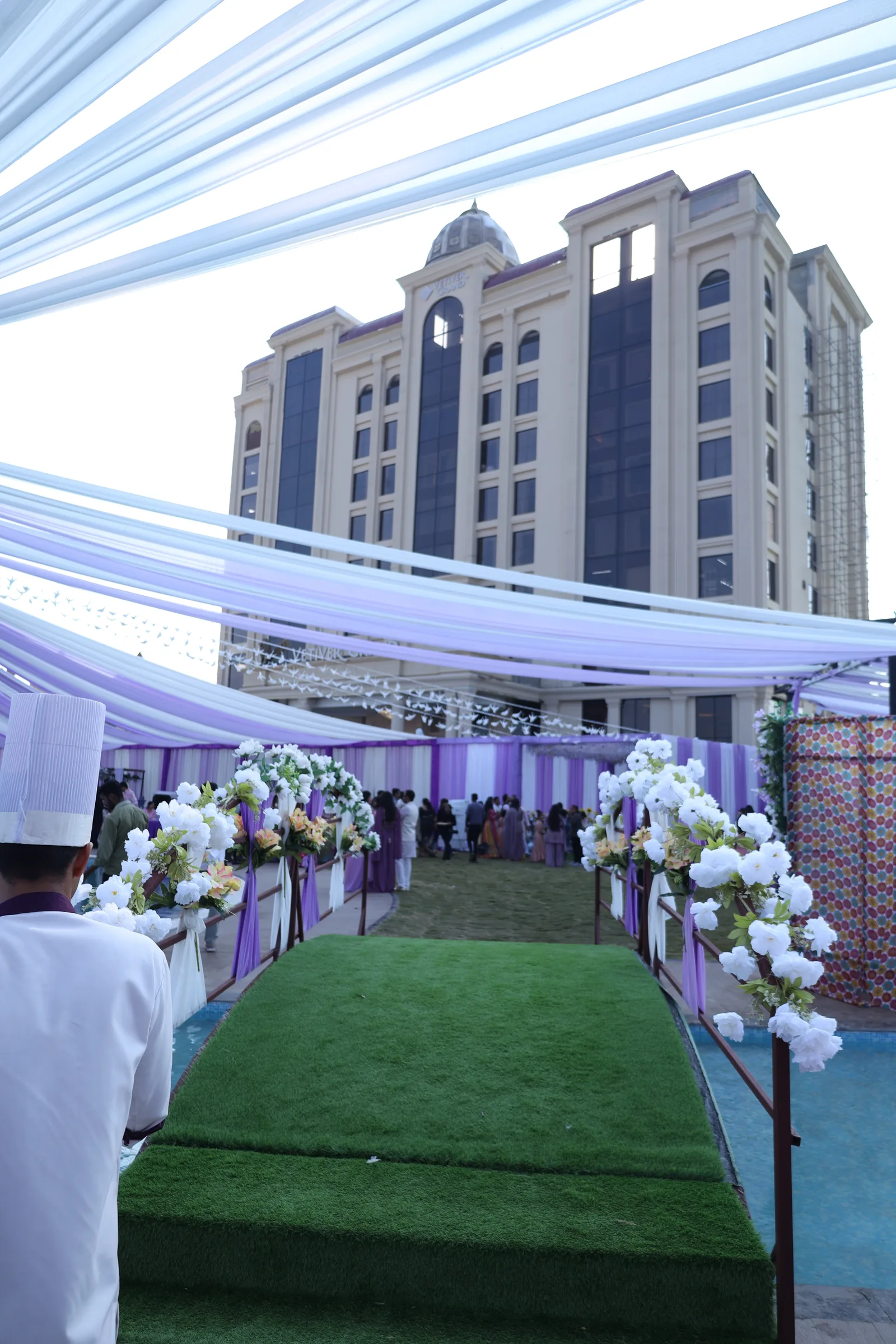Guests gather beneath lilac and white drapes at the end of a flower-lined walkway over the pool at Vetiver Grand, Jabalpur, during a daytime wedding celebration.