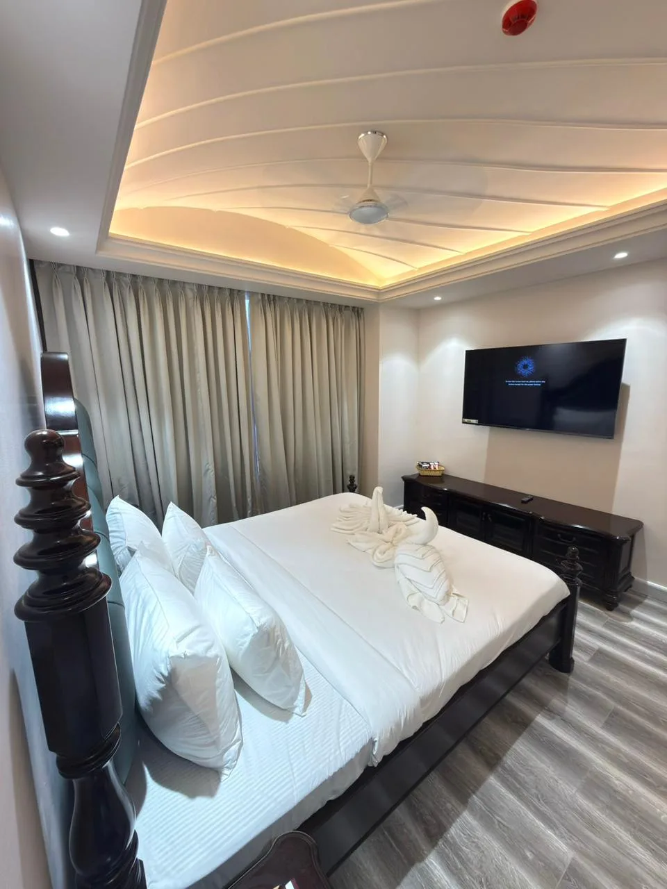 Pool-facing Executive room at Vetiver Grand, Jabalpur: dark carved four-poster bed dressed with swan towel art, facing the rooftop pool side of the house.